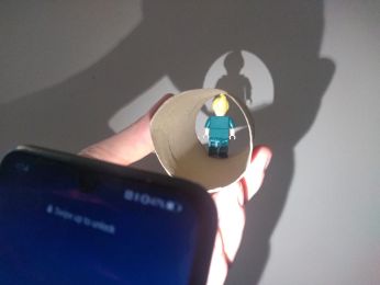 A phone torch, shinning a light on a LEGO minifigure man placed on the inside of a cardboard toilet roll